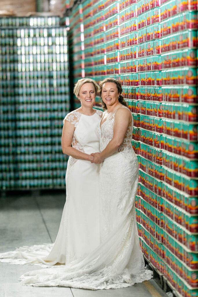 Two brides stand together in front of a colorful wall made by beer cans 
