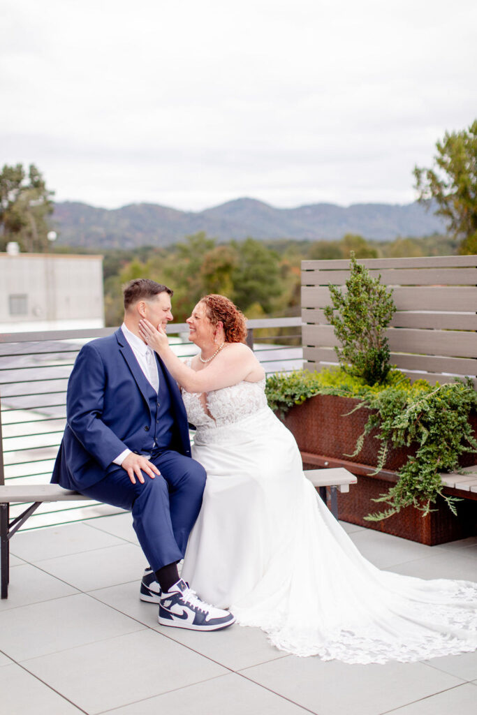 A wedding couple sit on a bench on a rooftop, sharing an intimate moment with a mountain view behind them. 