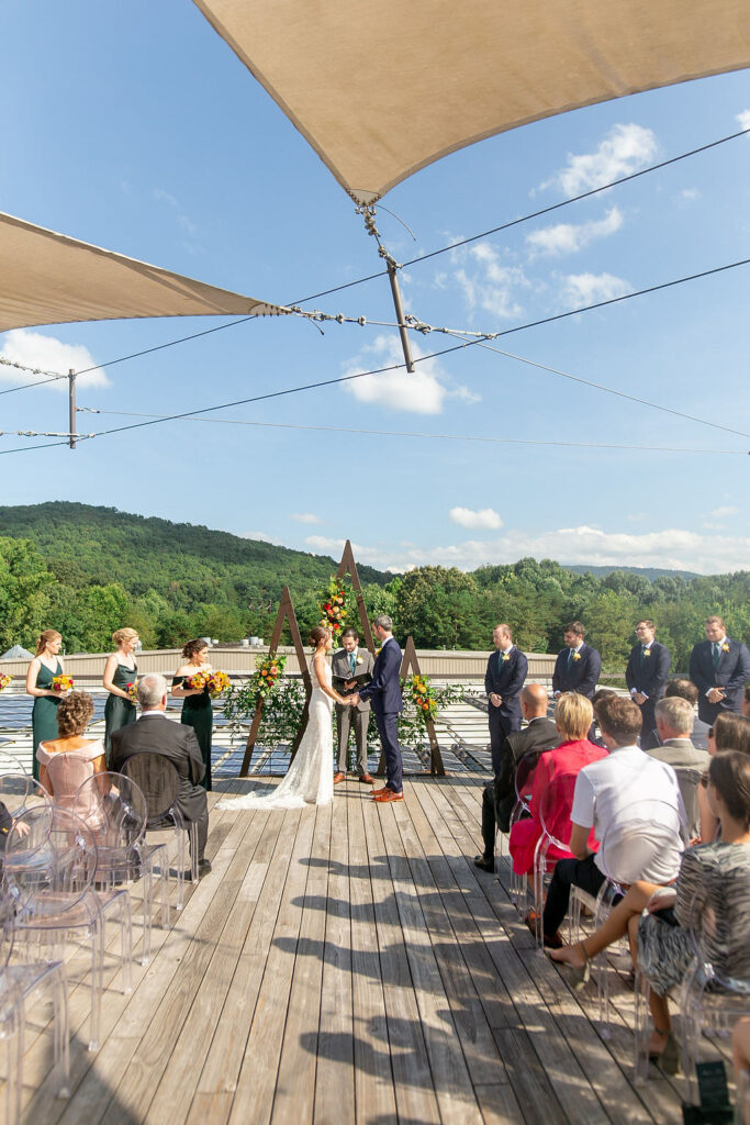 A wedding ceremony on a rooftop deck with mountains in the background. The couple stand under a triangular floral arch, surrounded by their wedding party and guests seated on either side