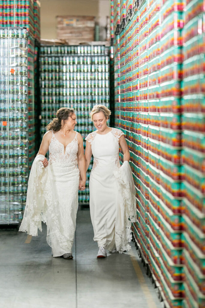 A wedding couple holding up the bottom of the dresses and walking along a wall of beer cans 