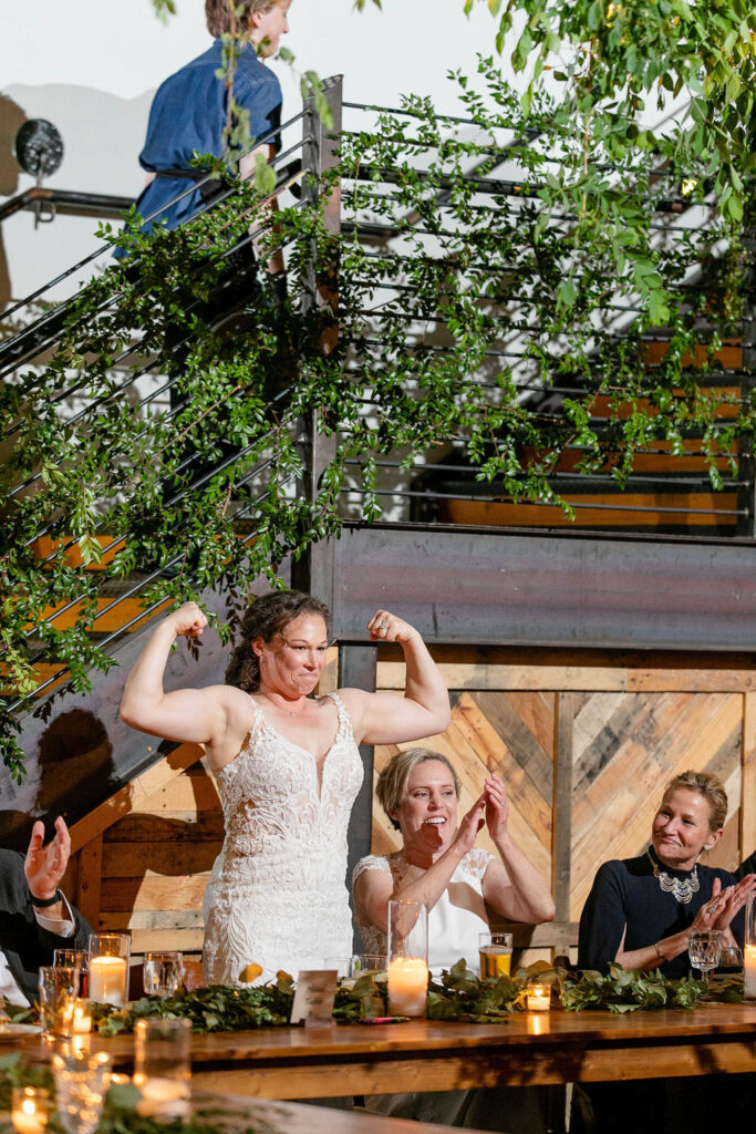 A bride standing up at a reception table flexing muscles 