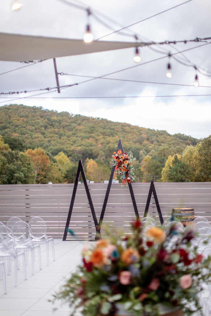 Clear chairs, flower arrangements, and wooden triangle arches set up for a wedding ceremony 