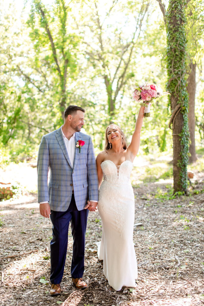 A newlywed couple walking in a wooded area together as one holds up a bouquet of flowers 