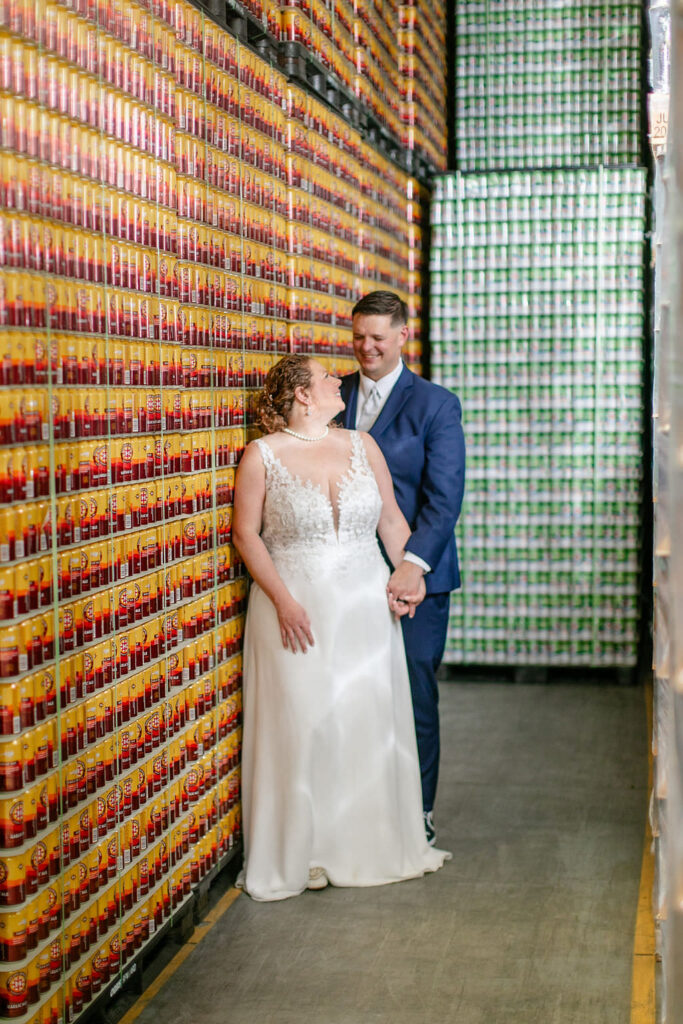 A newlywed couple standing together smiling in front of a beer wall. 