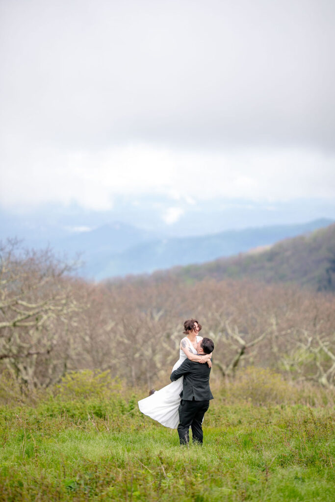 The groom lifting the bride up in a grassy field with bare trees and mountains in the background