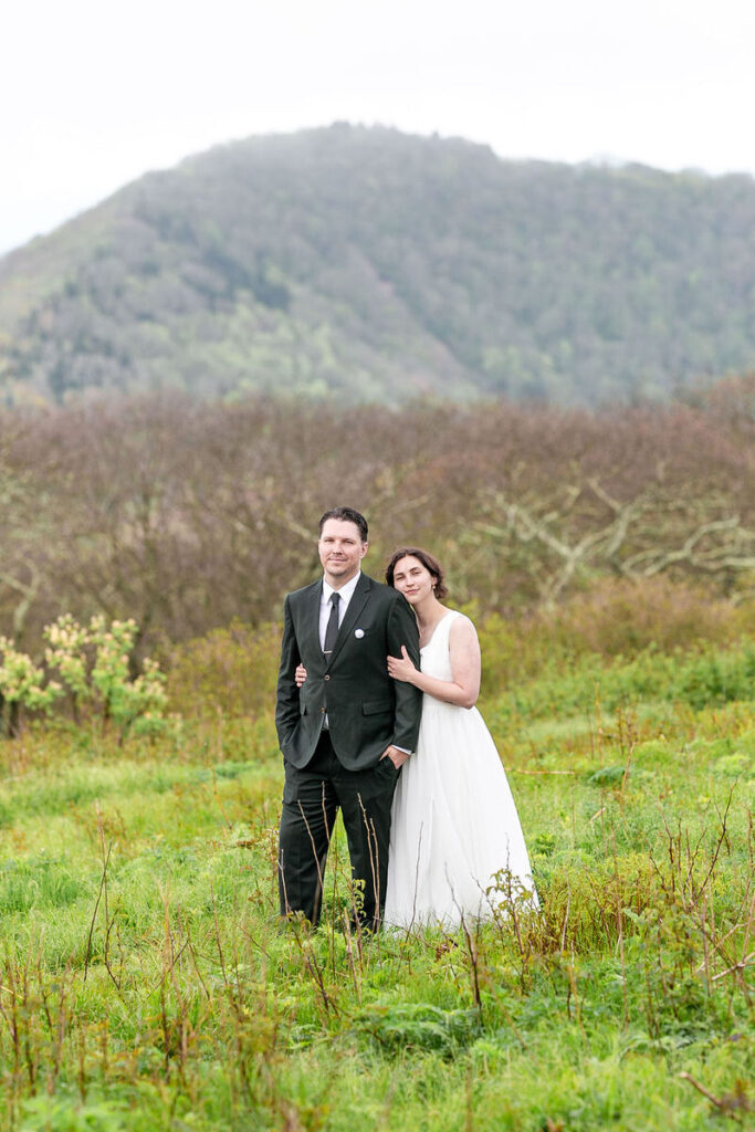 A couple standing in a green field with a mountainous background, the bride in a white dress and the groom in a black suit.