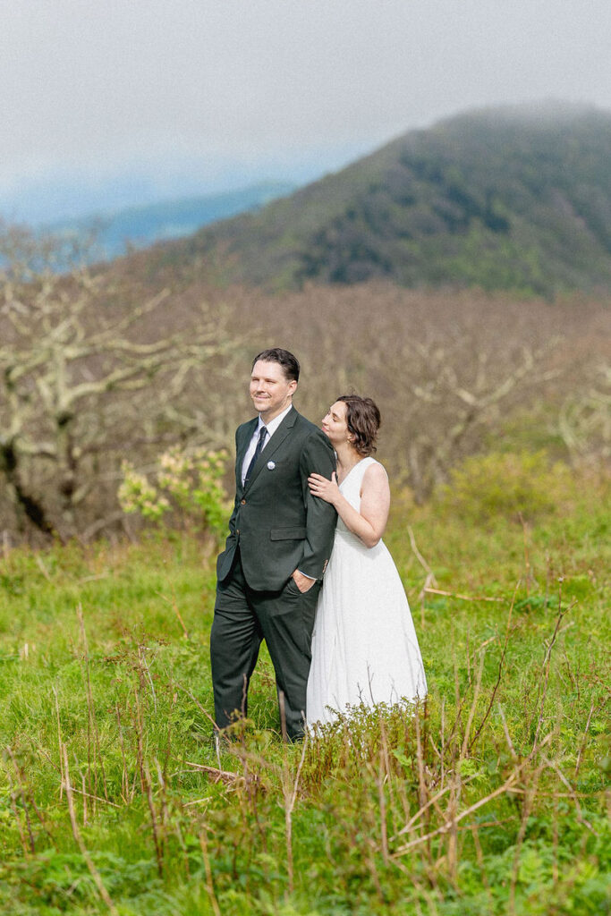 The bride and groom standing close together in a grassy field with mountains in the background. ​​