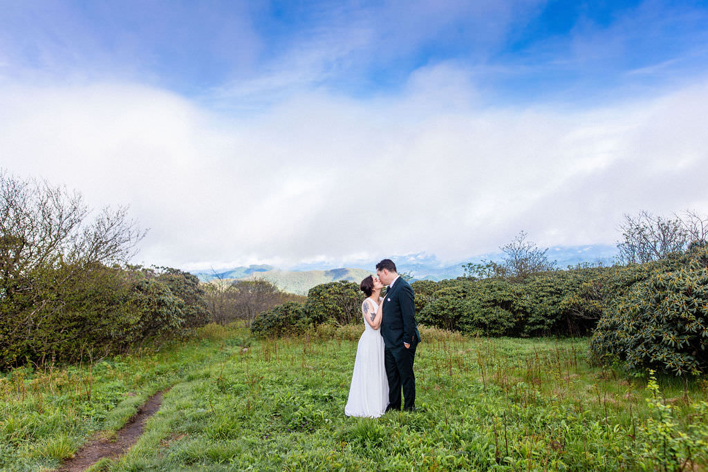 The bride and groom sharing a kiss in a green field with a vibrant blue sky and mountains in the background.
