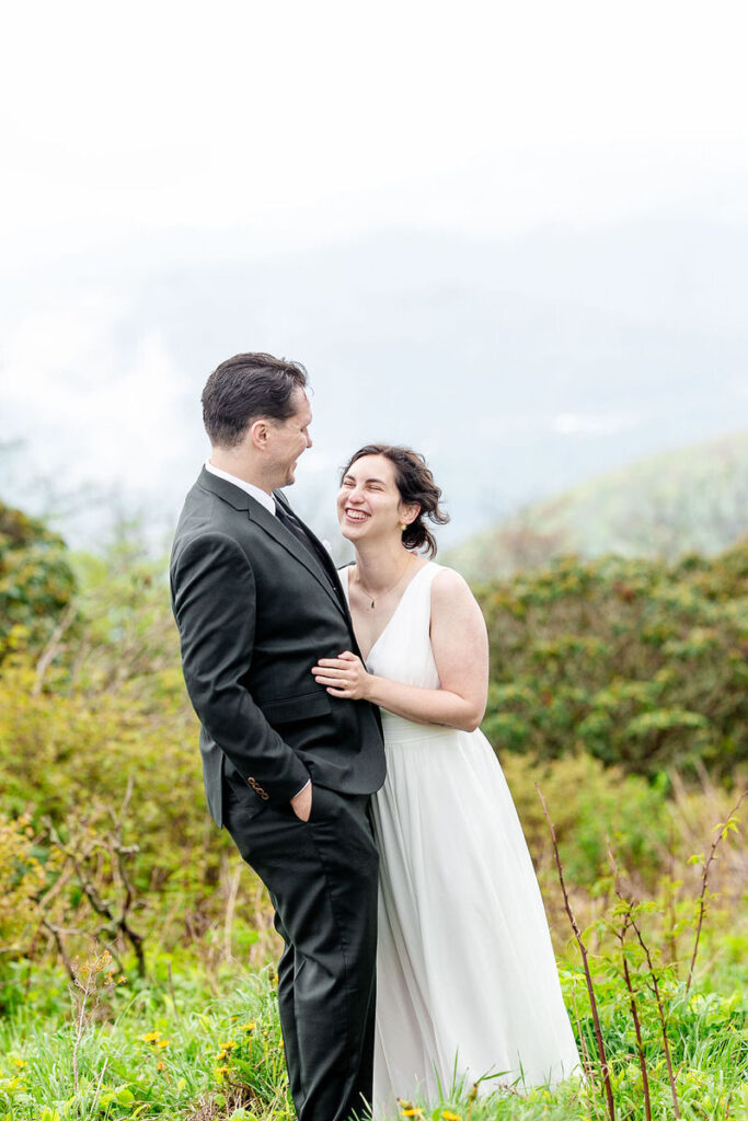 The bride and groom smiling and looking at each other in a green, scenic outdoor setting.
