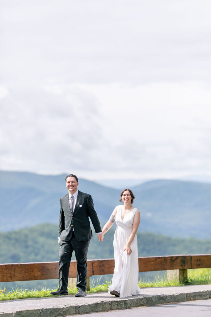 The bride and groom holding hands and walking on a roadside with expansive mountain views behind them