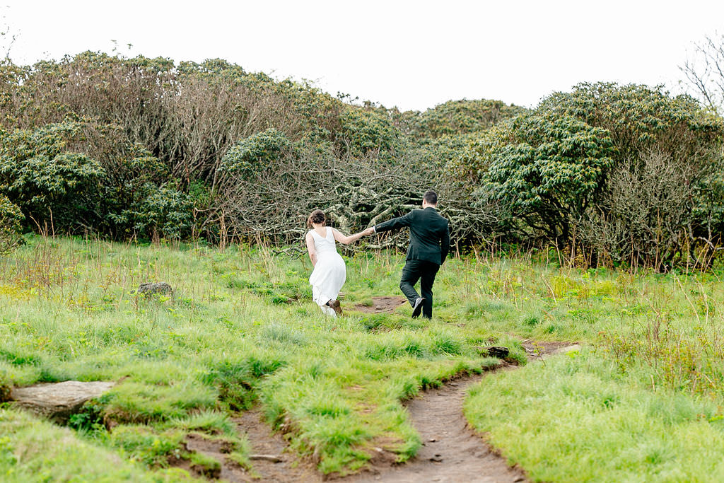 A wedding couple holding hands and walking on a dirt path in a grassy area 