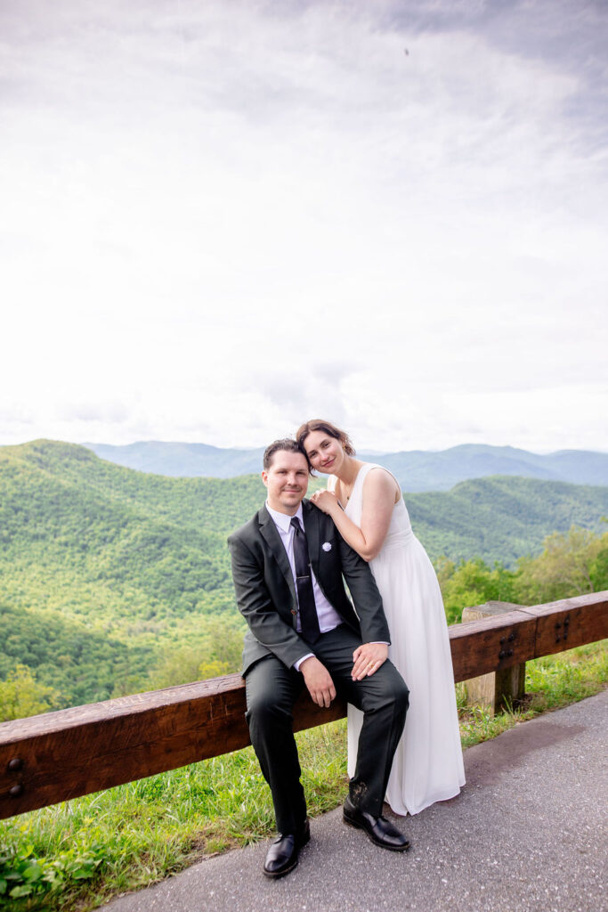 The bride and groom sitting on a wooden bench with a panoramic view of green mountains behind them