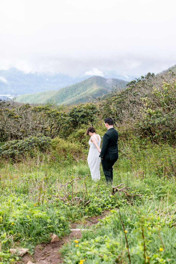 The bride and groom walking through a grassy path, with the bride holding the groom’s hand and looking down, surrounded by lush greenery and mountains