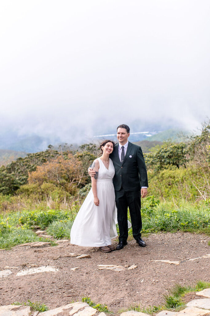 The bride and groom posing together in a mountain setting, smiling with mist-covered mountains in the background