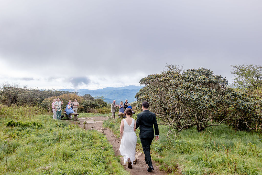 The bride and groom walking towards the wedding ceremony site, with guests waiting in the distance and a mountainous landscape behind them