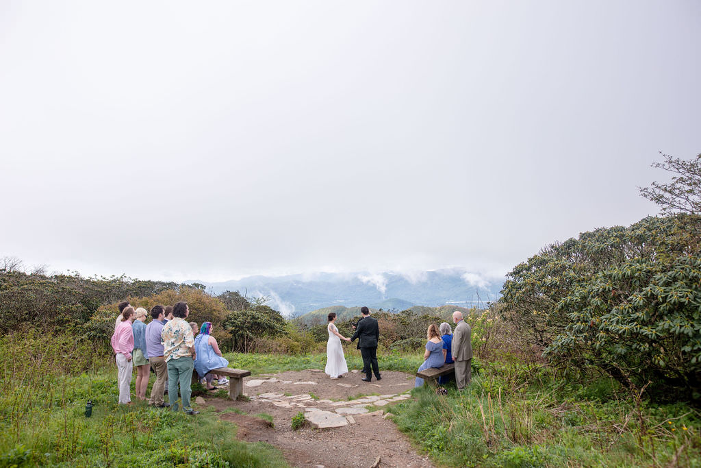 An outdoor wedding ceremony with guests gathered around a couple exchanging vows with a scenic mountainous view in the background