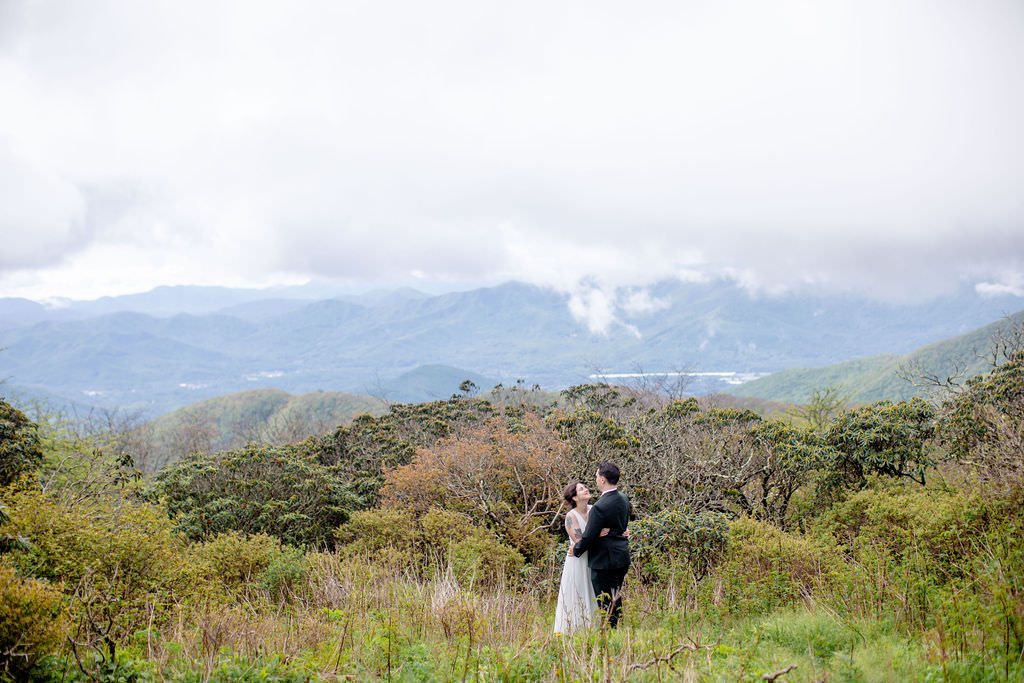 A newlywed couple standing in a grassy area overlooking the mountains with their arms around each other