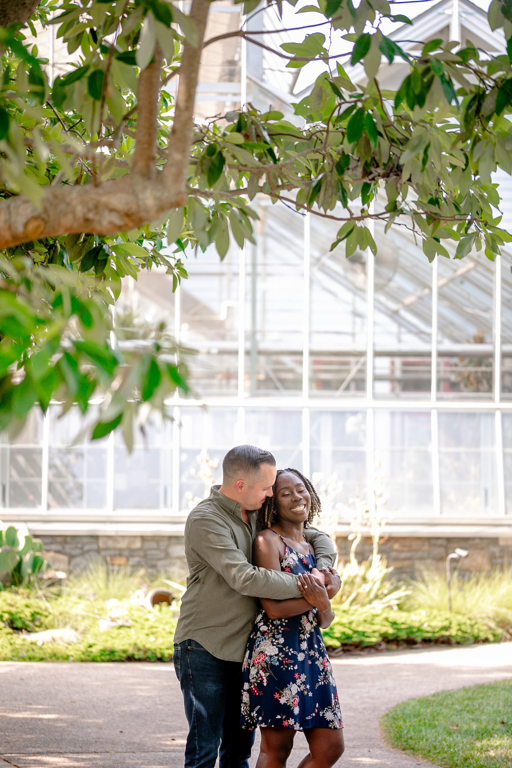 a man and a woman standing in front of a greenhouse - the man has his arms wrapped around the woman's shoulders 