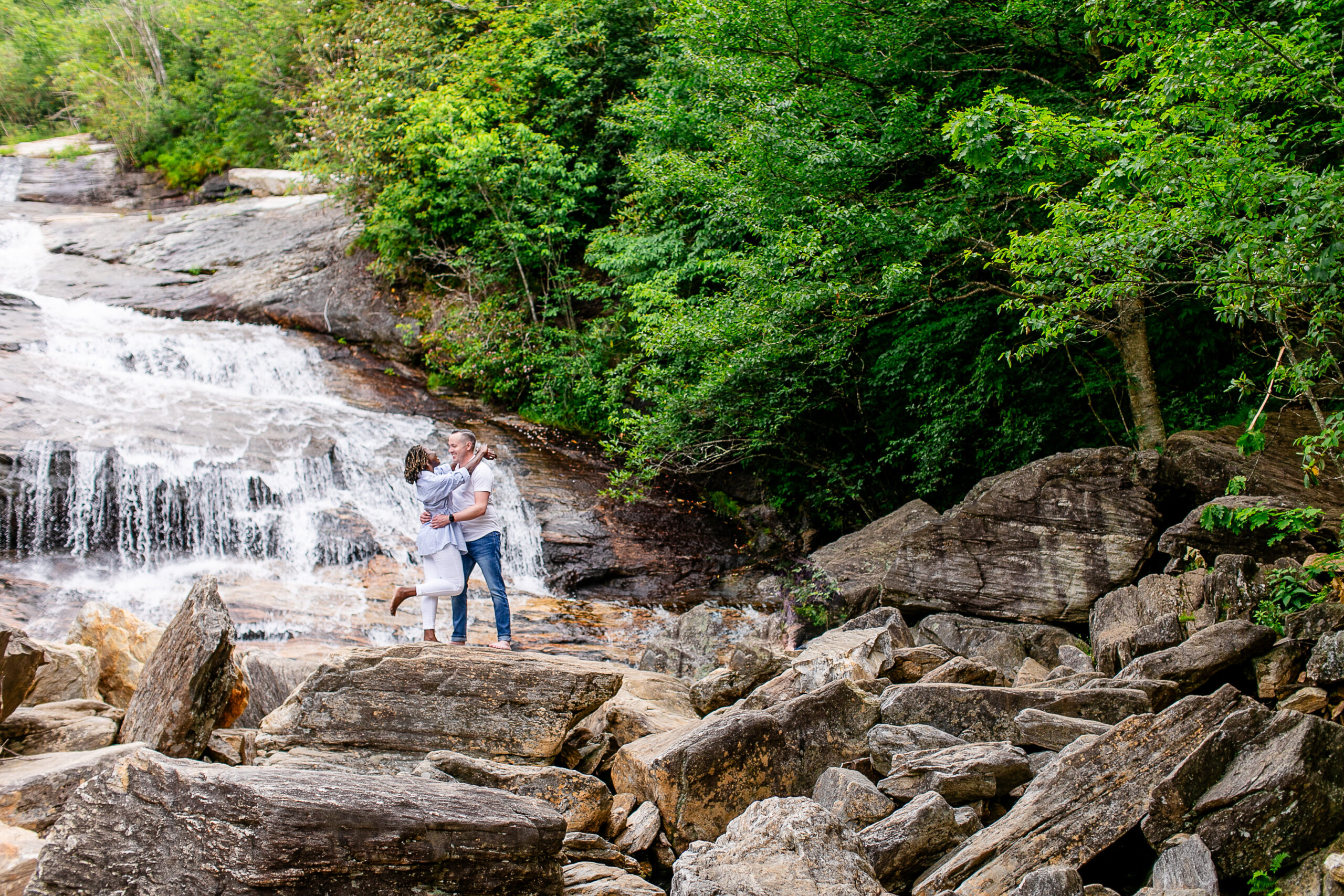 a man and woman holding each other with affection on rocks with a waterfall behind them and trees surrounding them 