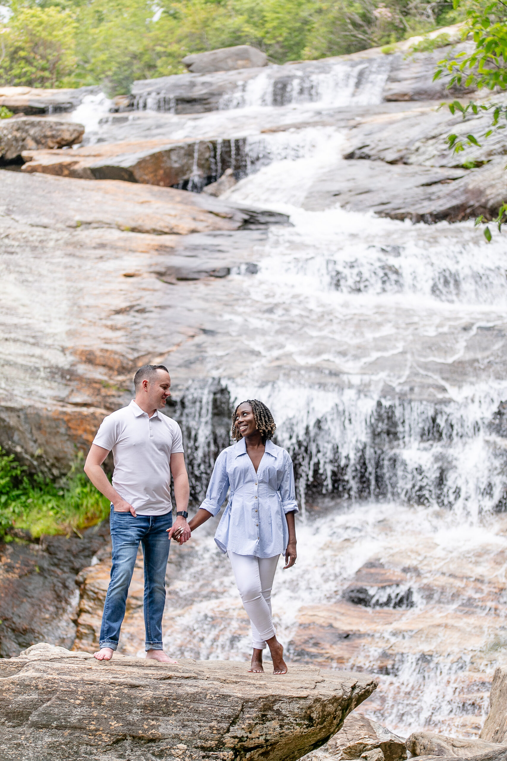 a man and a woman looking at each other with a waterfall as their backdrop, both are holding hands 