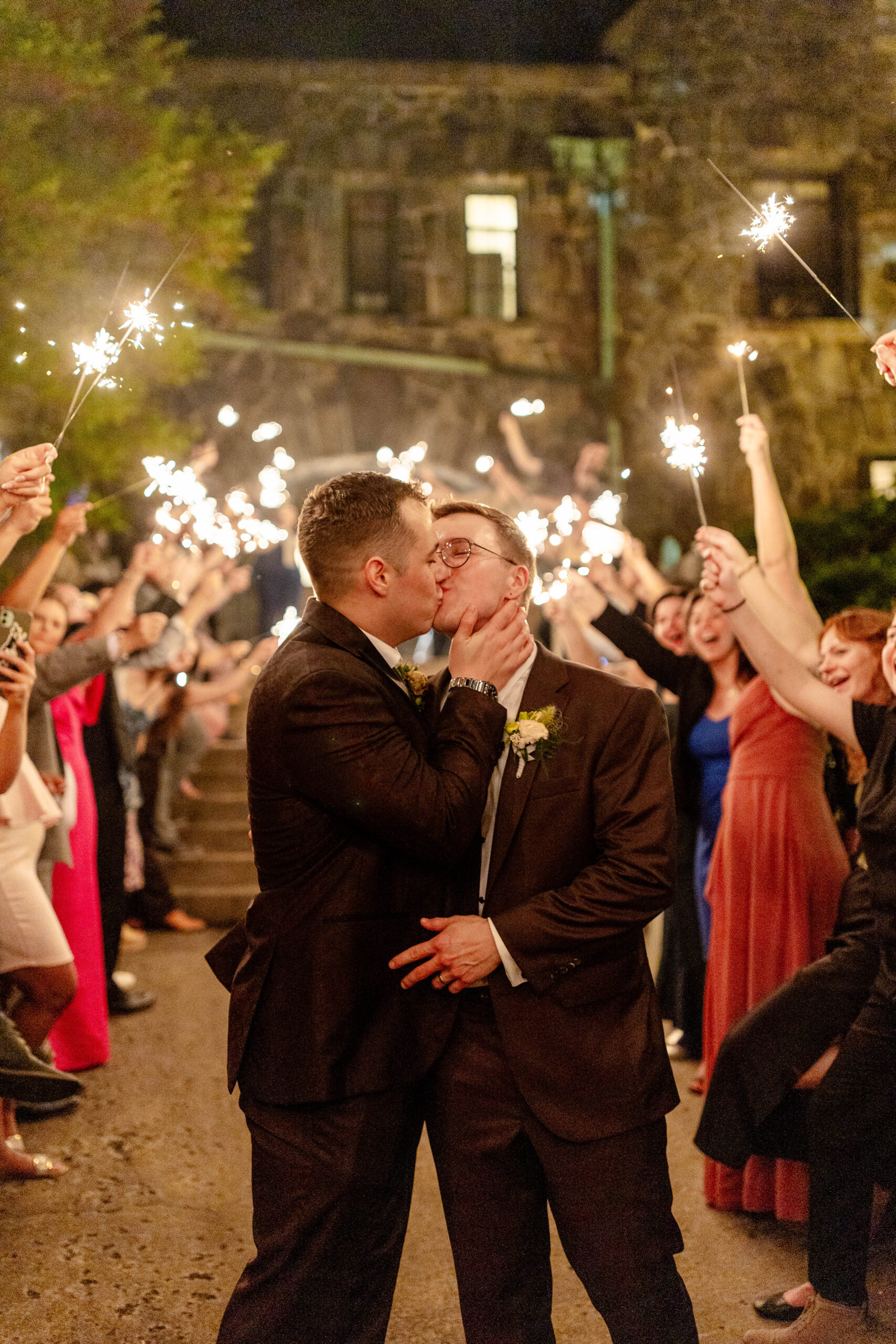 two men who just got married giving each other a kiss surrounded by their wedding guests holding sparklers