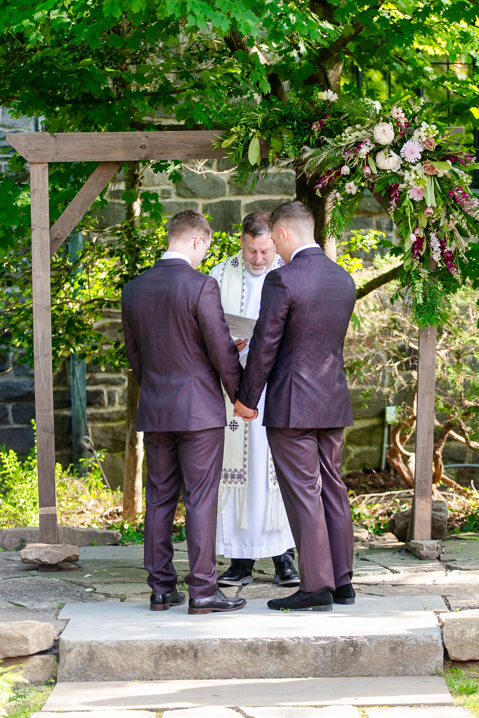 two men about to get married holding hands under a wooden arch in front of their wedding officiant 