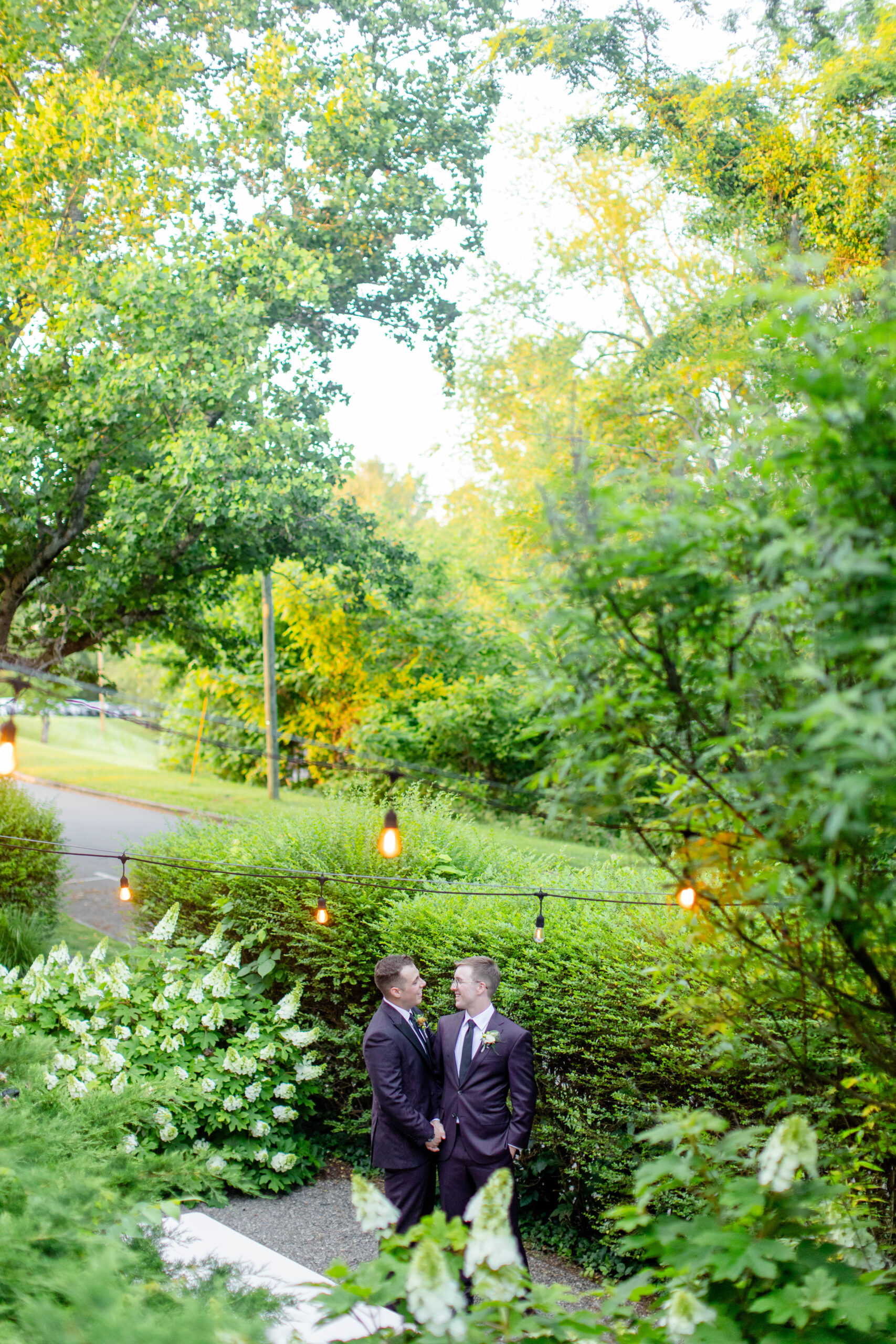 two men in suits about to be married looking at each other surrounded by trees and lights 