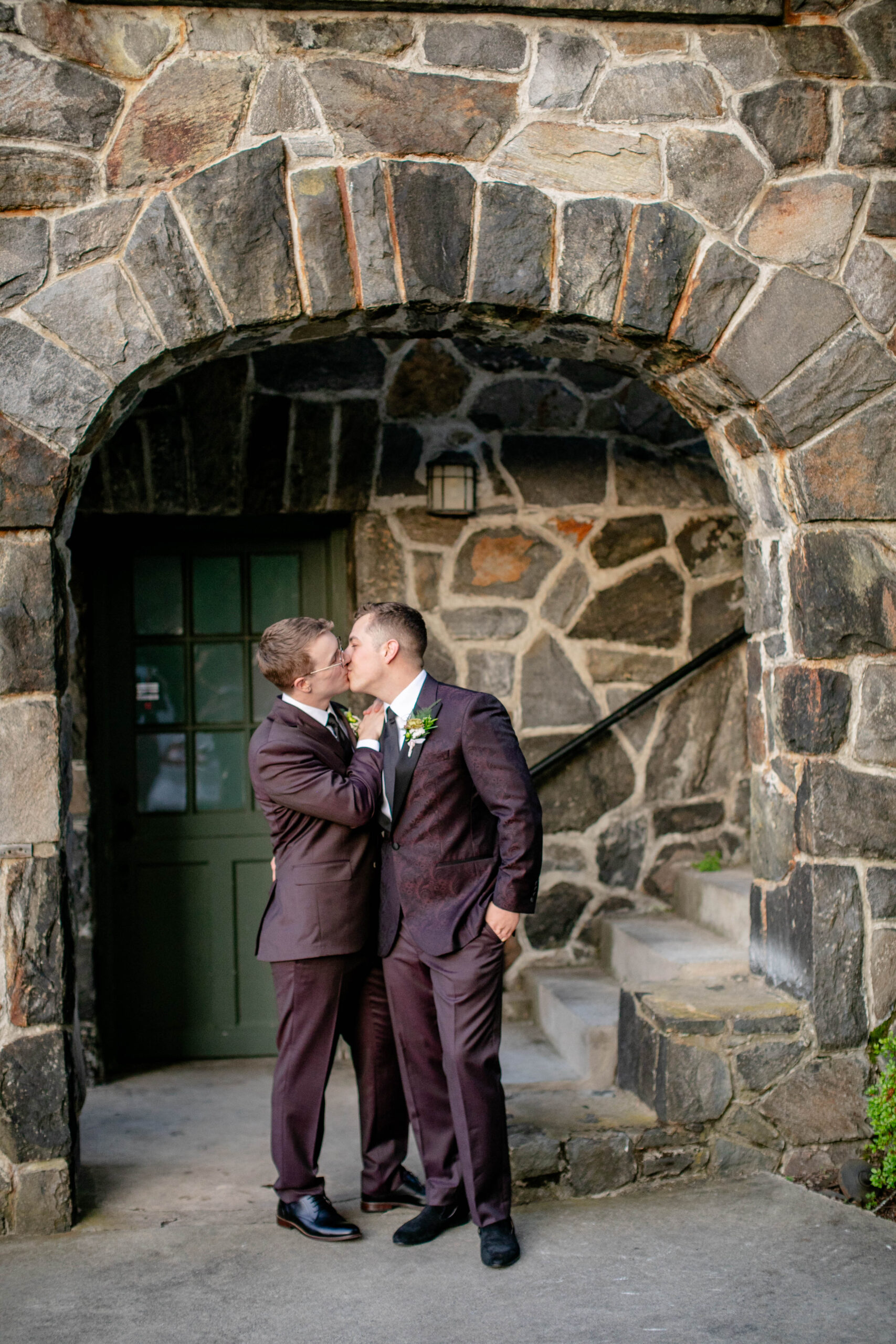 two men giving each other a kiss in their suits underneath a stone archway 