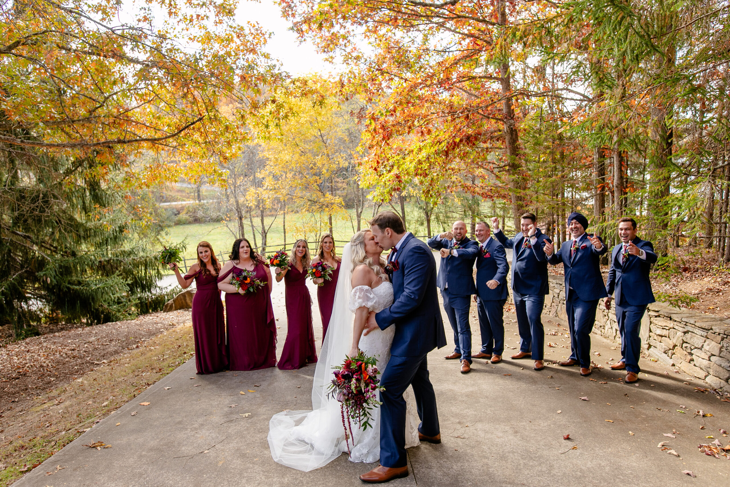 bride and groom kissing with wedding party behind them surrounded by fall folliage