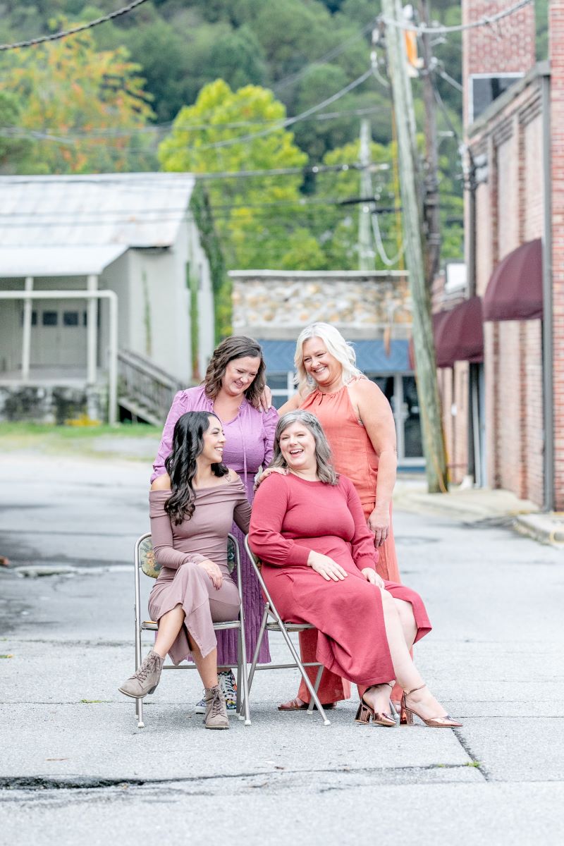 a group of women posing for a picture two are sitting on folding chairs and two women are standing behind them all the women are laughing together 