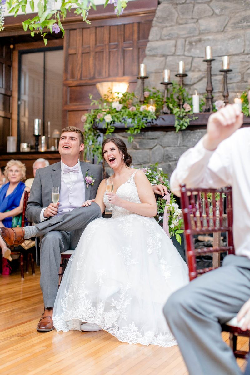 a man and woman sitting in chairs and laughing the woman is wearing a white wedding dress and holding a champagne glass the man is wearing a gray suit and also holding a champagne glass