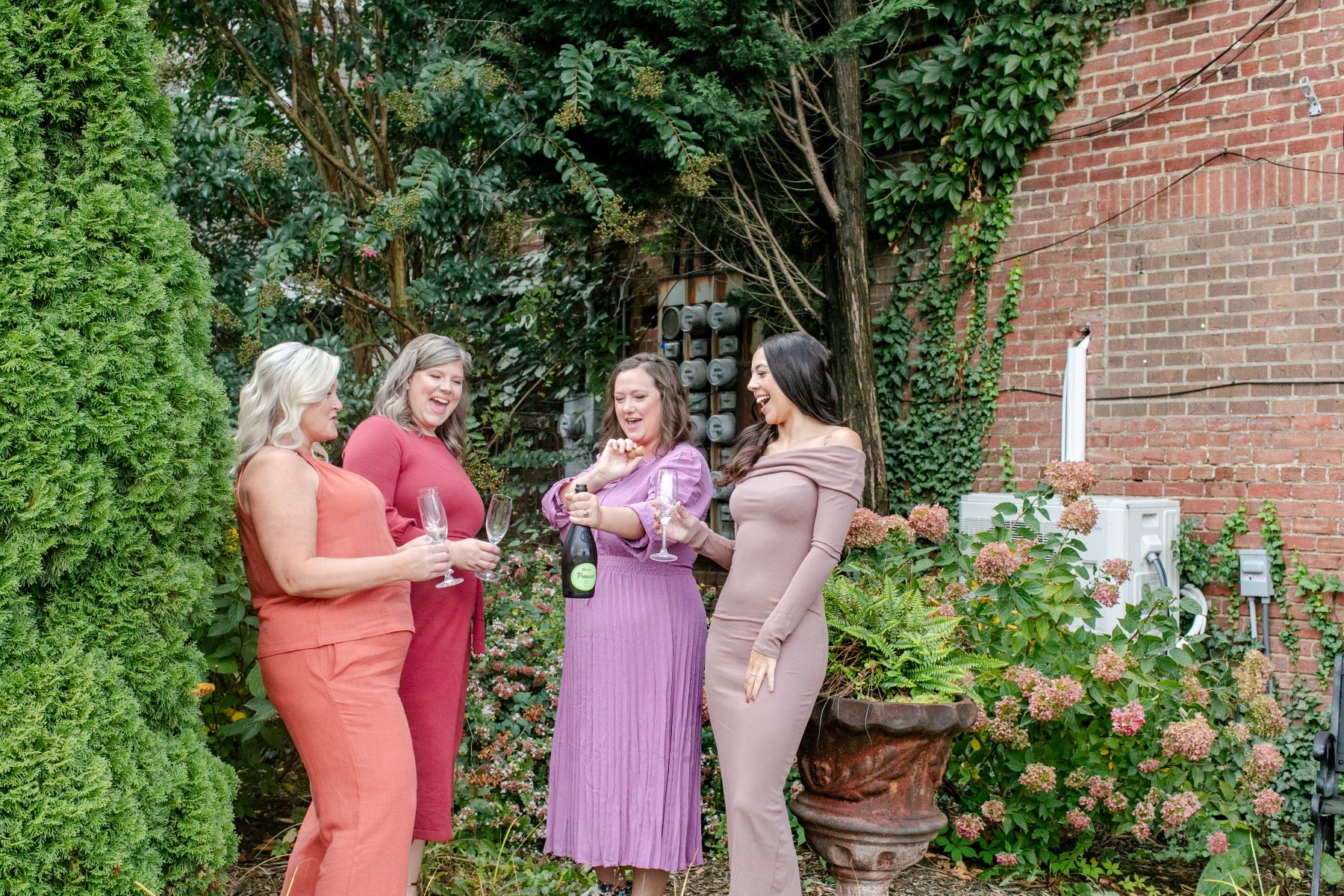 a group of women in dresses standing in a yard one woman is popping a bottle of champagne andn the other women are holding champagne glasses 