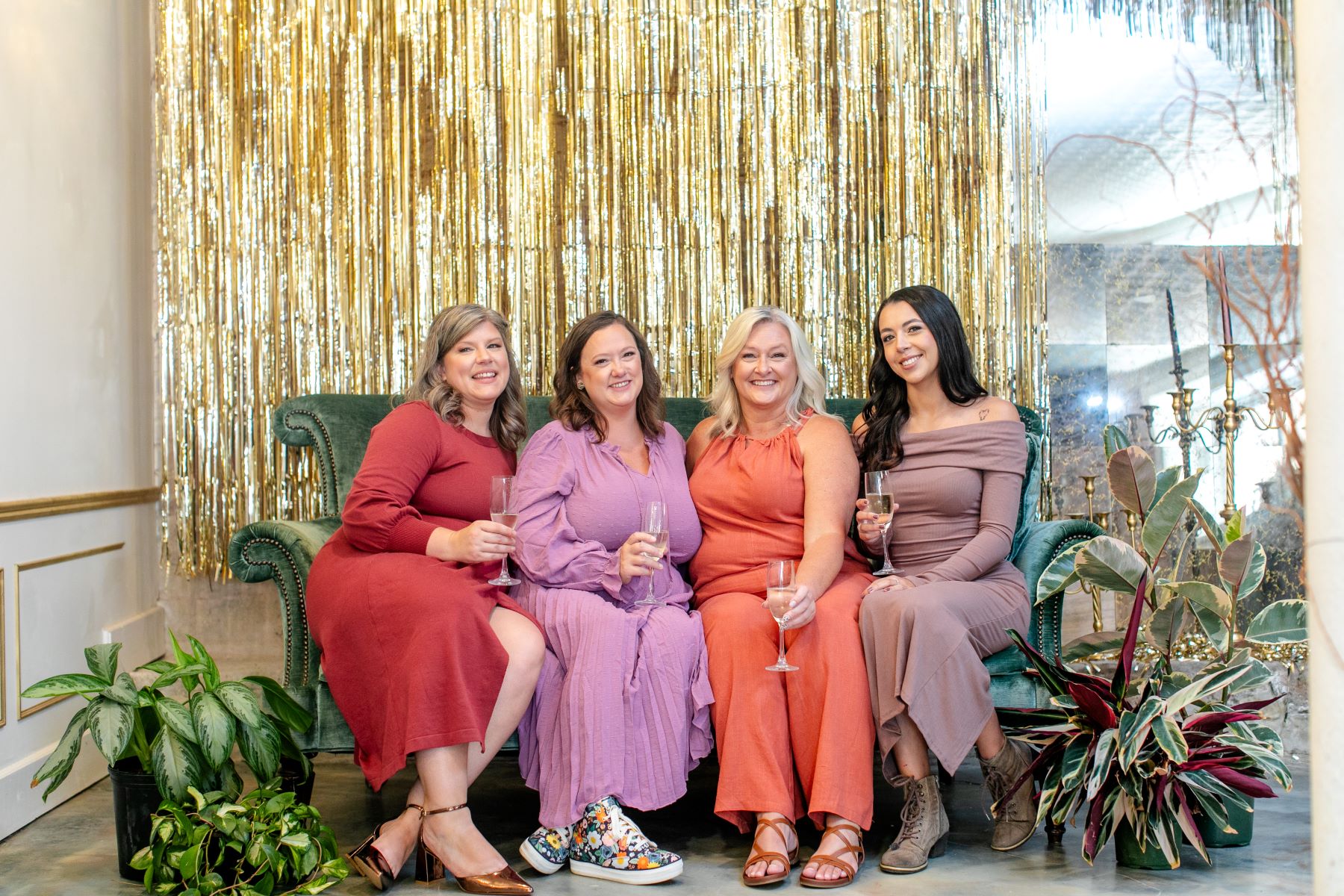 four women sitting together on a green velvet couch all wearing dresses and holding champagne glasses and smiling