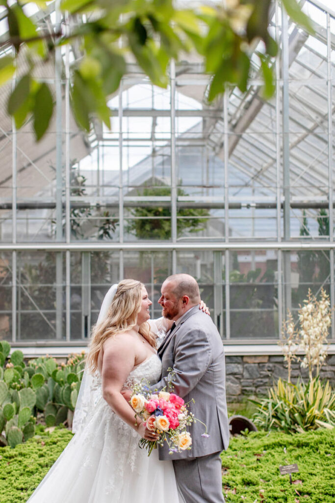 Newlyweds with their arms around each other standing outside of a greenhouse 