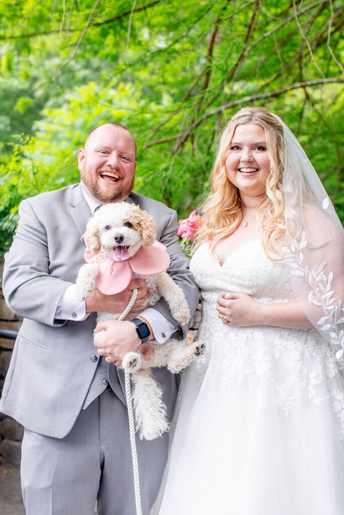 Two people in wedding outfits smiling as one of them holds up a small dog. 