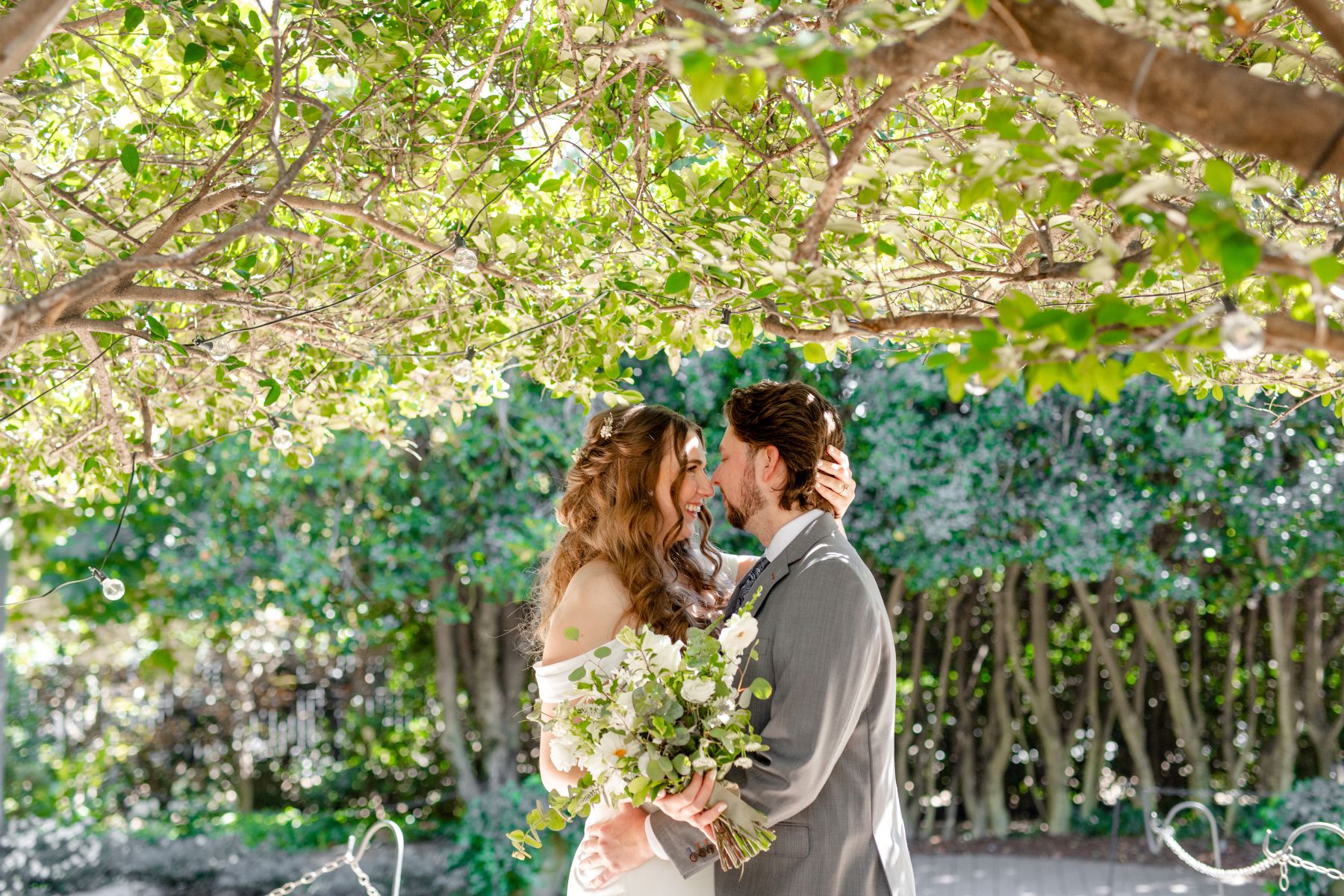 A woman in a white wedding dress holding a bouquet of white flowers with her other hand on the back of her partner's head smiling at him her partner is wearing a gray suit and has his hand on her waist behind them is a grove of trees 