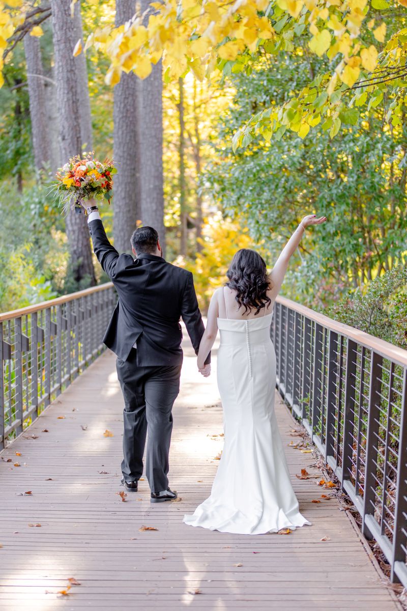 A couple walking on a bridge together with their hands up in the air and also holding hands the woman is wearing a white dress the man is wearing a black suit and is holding up a bouquet of flowers 