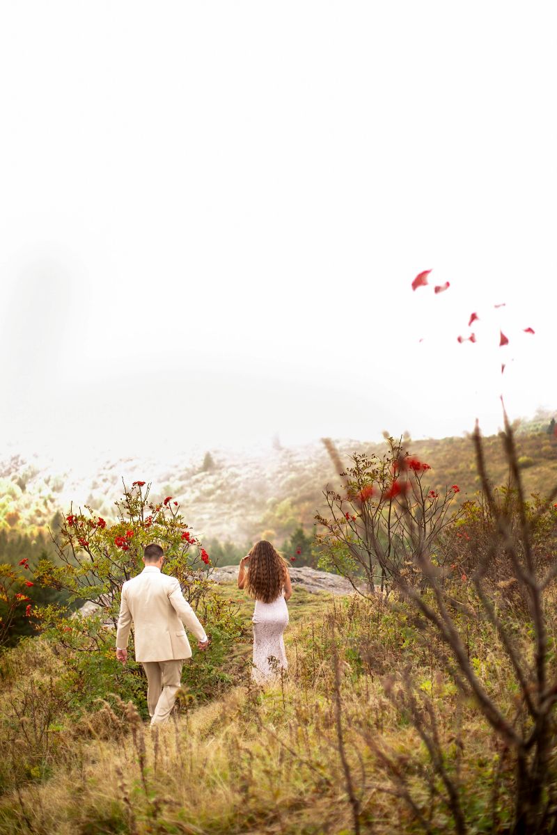 A couple walking through a field on a grassy mountain top the woman is wearing a white dress the man is wearing a tan suit 