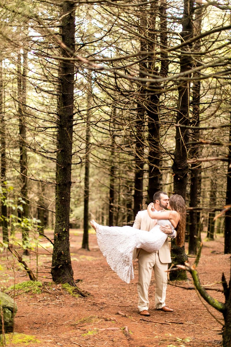 A couple in a forest surrounded by pine tress the man is lifting up his partner in the air and kissing her the man is wearing a beige suit the woman is wearing a white wedding dress 