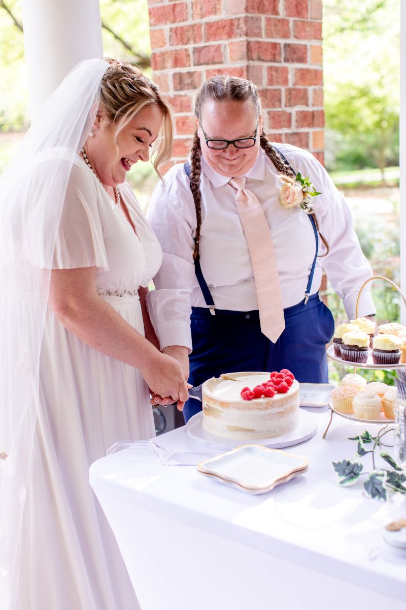 A couple cutting their wedding cake one is wearing a white wedding dress and white veil and their partner is wearing a white button up , a peach colored tie and blue suspenders they are both smiling 