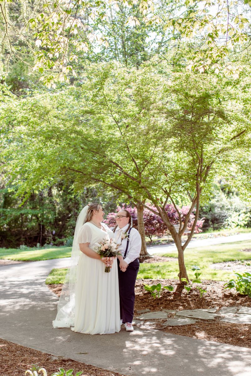 A couple standing on a sidewalk underneath a grove of treees smiling at each other and holding hands one is wearing a white wedding dress, a white veil and holding a bouquet of folowers the other is wearing a button up and suspenders 