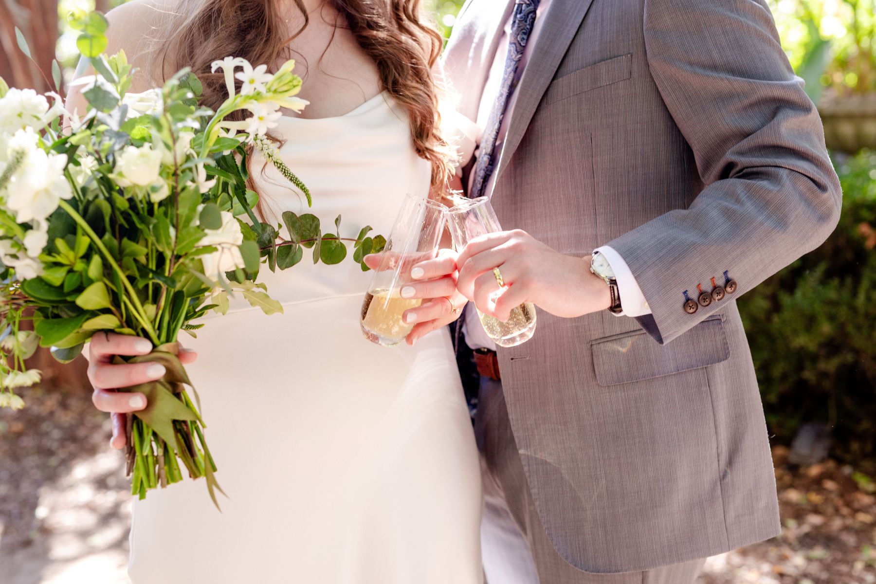 A couple clinking their glasses with champagne together the woman is wearing a white wedding dress and holding a bouquet of flowers the man is wearing a gray suit 