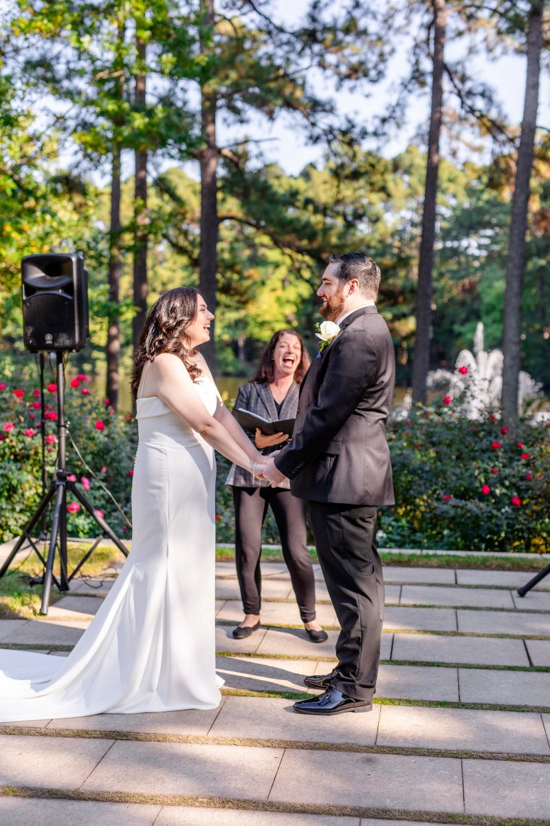 A couple at their elopment ceremony with their officiant holding hands and smiling at one another the woman is wearing a white dress the man is wearing a black suit 