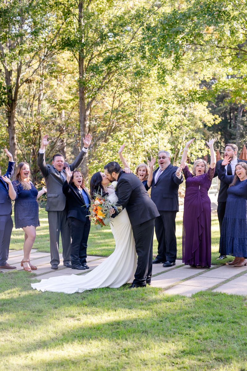 A couple kissing after their wedding ceremony and their guests are cheering them on standing behind them the woman is wearing a white dress and holding a bouquet of flowers the man is weraing a black suit 