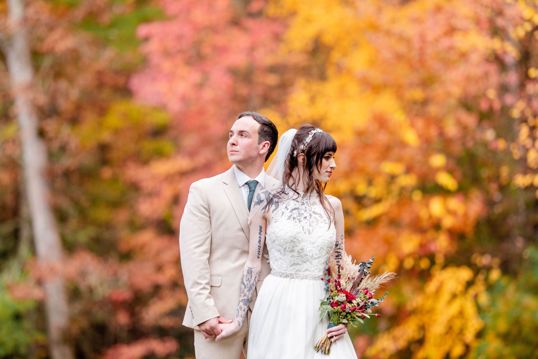 a couple standing together in front of fall foliage the man is looking to one side the woman is looking to the other the woman is wearing a white wedding dress and holding a bouquet of flowers and has on a white veil she also has tattoos on her arms the man is wearing a tan suit 