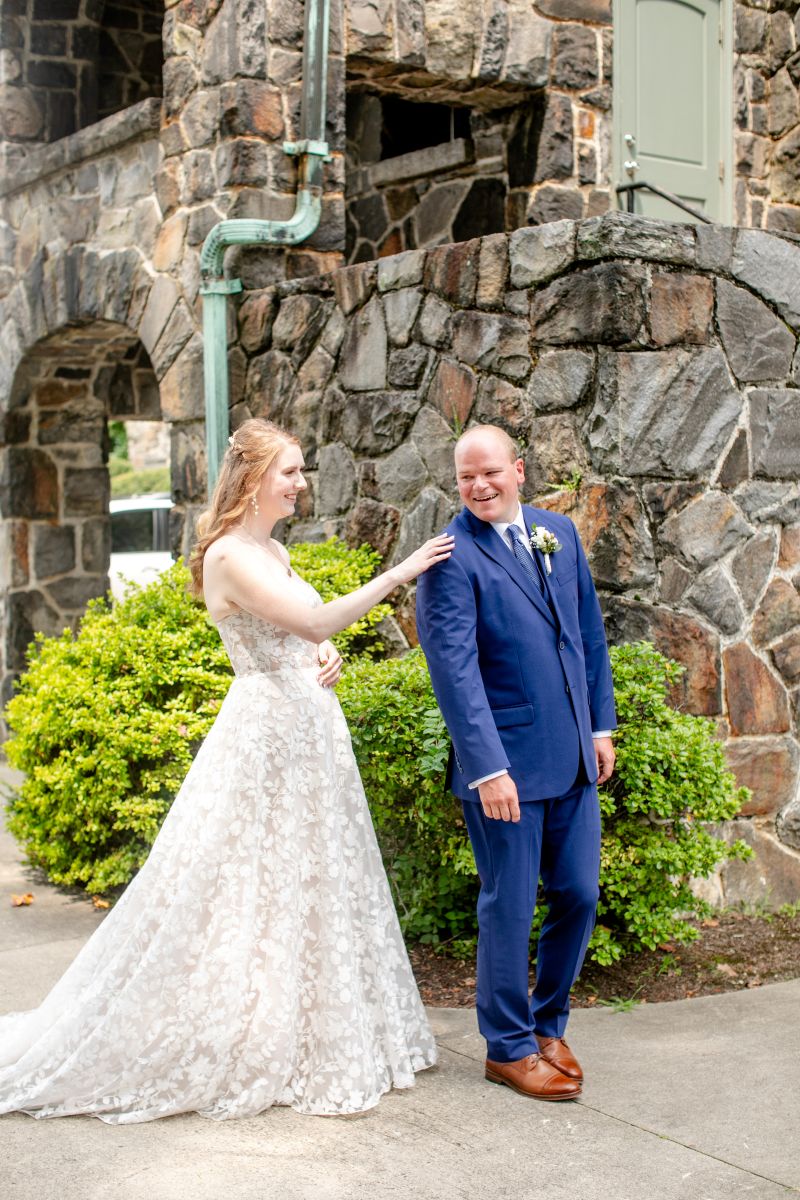 a couple having a first look together the woman is wearing a white wedding dress the man is waering a blue suit the woman is touching the man on the shoulder 