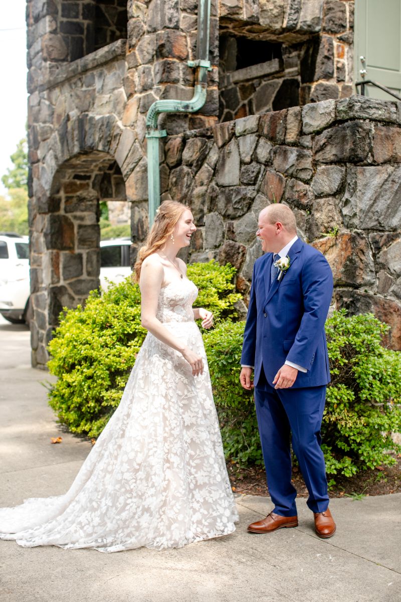 a couple sharing a first look together and laughing with one another the woman is wearing a white wedding dress the man is wearing a blue suit 