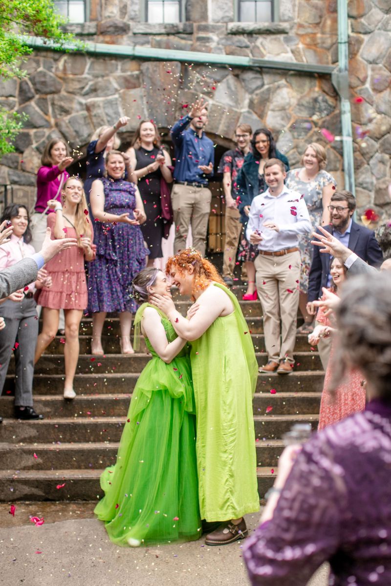 a couple both wearing green dresses holding each other in front of a staircase and laughing behind them are their wedding guests who are cheering and throwing confetti 