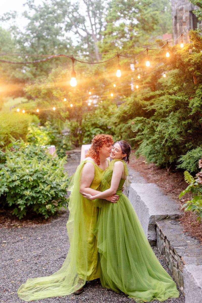 a couple both wearing green dresses standing underneath twinkling lights and hugging one another one partner is kissing the other on the cheek 