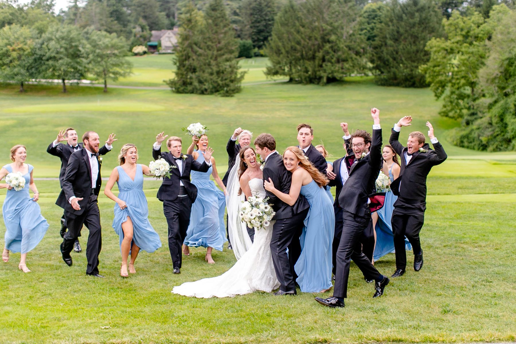 a couple on a golf course laughing together the woman is wearing a white wedding dress and veil and holding a bouquet of flowers the man is wearing a black suit and their wedding guests are surrounding them and cheering for them